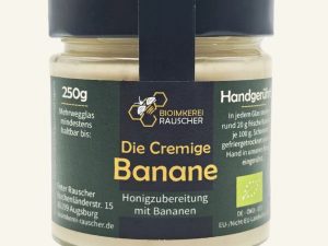 Vorderansicht des 250g-Glases der Bananen-Honigzubereitung der BioImkerei Rauscher mit zentralem schwarzem Etikett, goldenem Schriftzug und sanft pastellgelben Seitenbalken. Im transparenten Glas ist die helle, cremige Masse sichtbar.