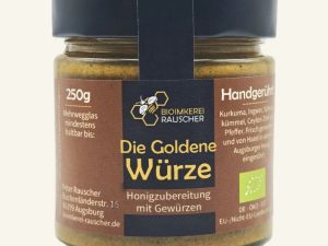 Vorderansicht des 250g-Glases „Die Goldene Würze“ der BioImkerei Rauscher. Das Etikett hat ein zentrales schwarzes Feld mit goldenem Bienen-Logo und großem goldenem Schriftzug „Die Goldene Würze“, eingefasst von Seitenbalken in mattem Ziegelrot. Der Inhalt ist ein leuchtend gelb-oranger Honig-Gewürz-Aufstrich.