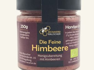 Vorderansicht des 250g-Glases „Die Feine Himbeere“ der BioImkerei Rauscher mit zentralem schwarzem Etikett, goldenem Schriftzug und pastell-altrosafarbenen Seitenbalken. Im transparenten Glas ist der fruchtig-rosafarbene Himbeer-Honigaufstrich zu sehen.